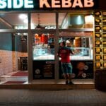 Nocna fasada kebabowni z podświetlonym menu i stojącym klientem