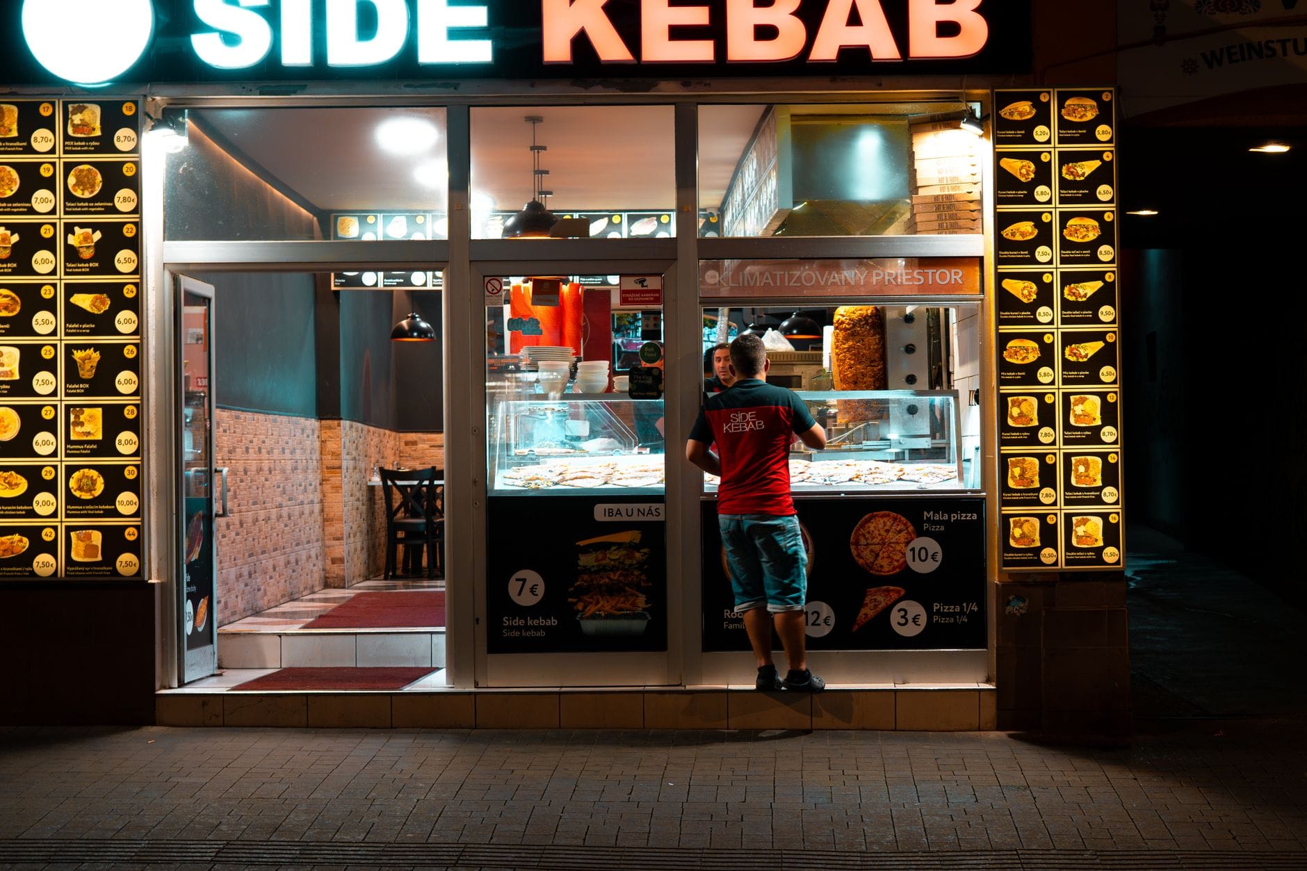 Nocna fasada kebabowni z podświetlonym menu i stojącym klientem